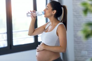 Pretty young pregnant woman drinking water while standing at home.
