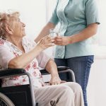 Professional nurse in a blue uniform gives a glass of water to an elderly woman in a wheelchair