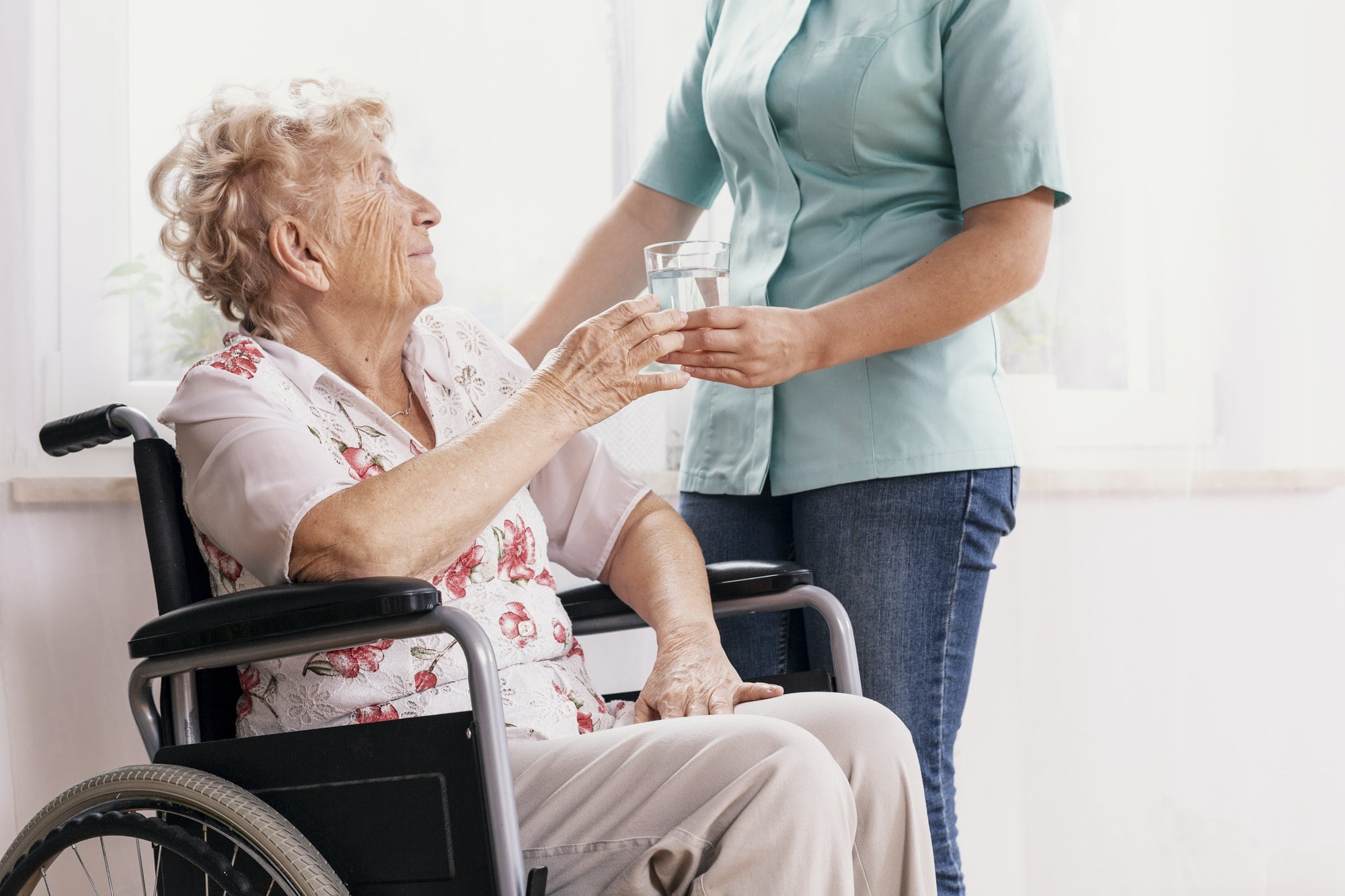 Professional nurse in a blue uniform gives a glass of water to an elderly woman in a wheelchair